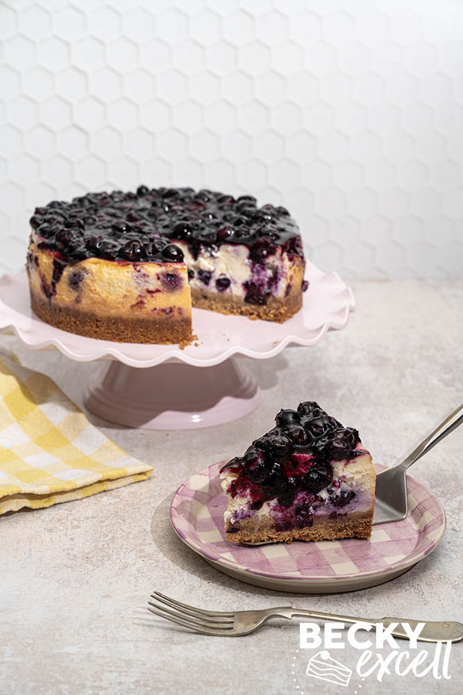 Both the full baked blueberry cheesecake on a cake stand and a slice placed on a serving plate
