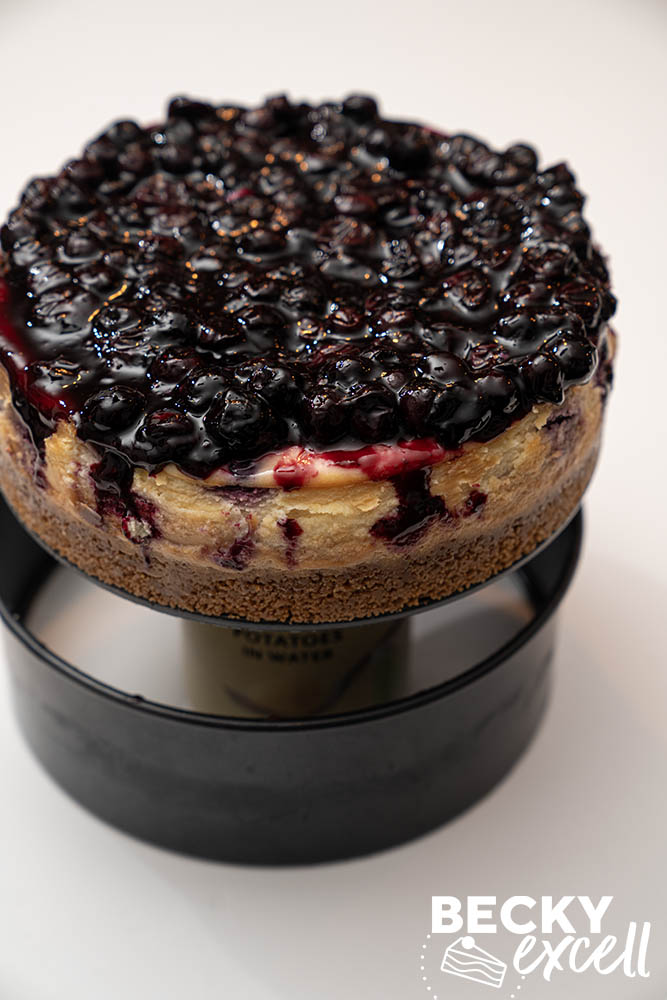 A clever kitchen hack demonstrated to remove a baked cheesecake from a loose-bottomed tin: rest it on a can and pull the sides down firmly and gently
