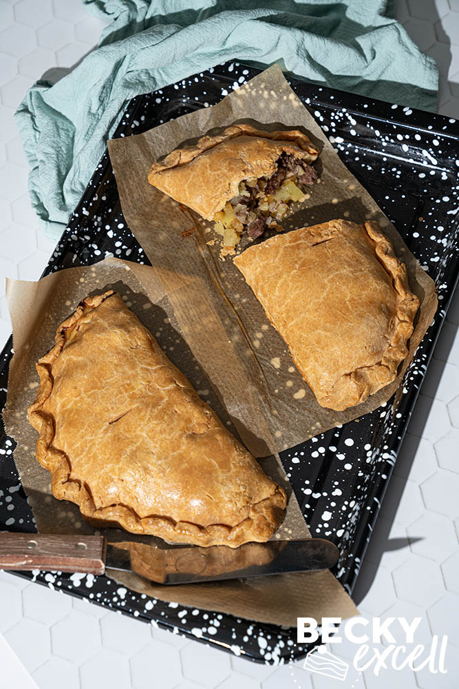 Two homemade gluten-free Cornish pasties on baking paper, one whole and one sliced in half