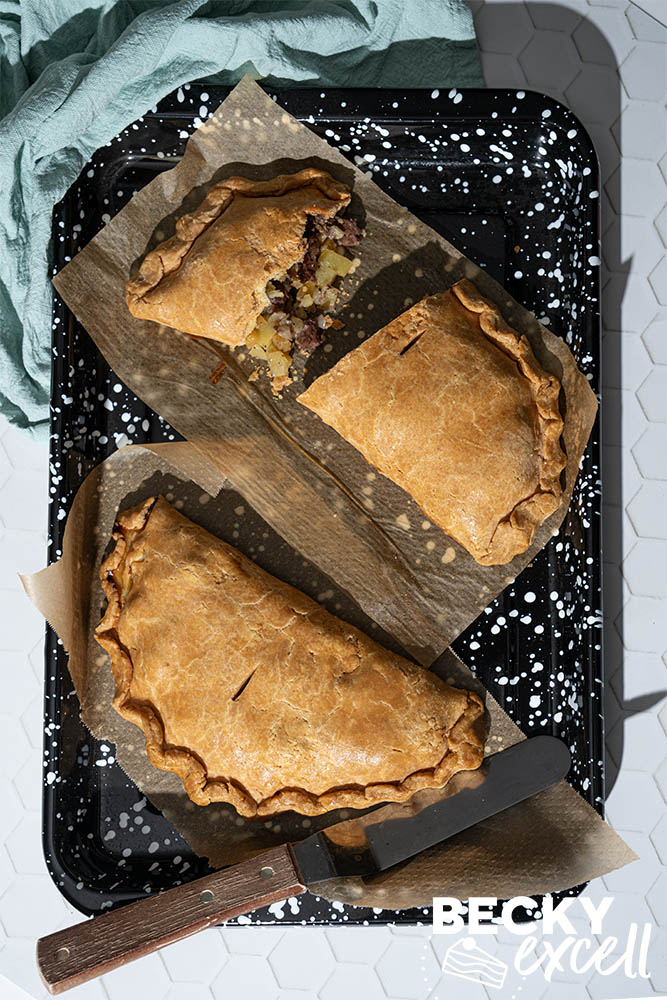 Two golden gluten-free Cornish pasties on a baking tray, with one cut open to show the beef and vegetable filling